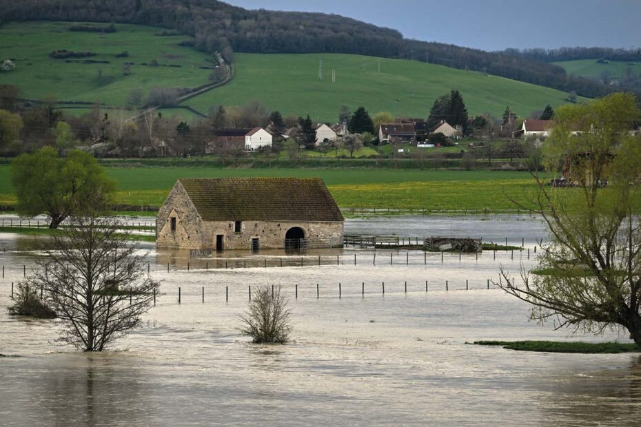 Bâtiment en pierre entouré par les eaux d’une inondation, avec des arbres immergés et un village en arrière-plan au pied des collines.