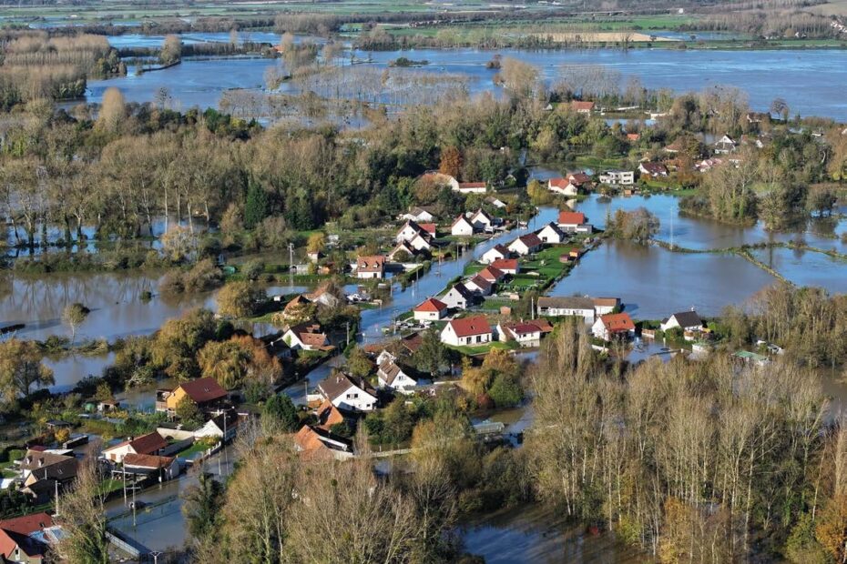 Vue aérienne d’un quartier résidentiel touché par une inondation, avec des maisons entourées d’eau et des zones boisées submergées.