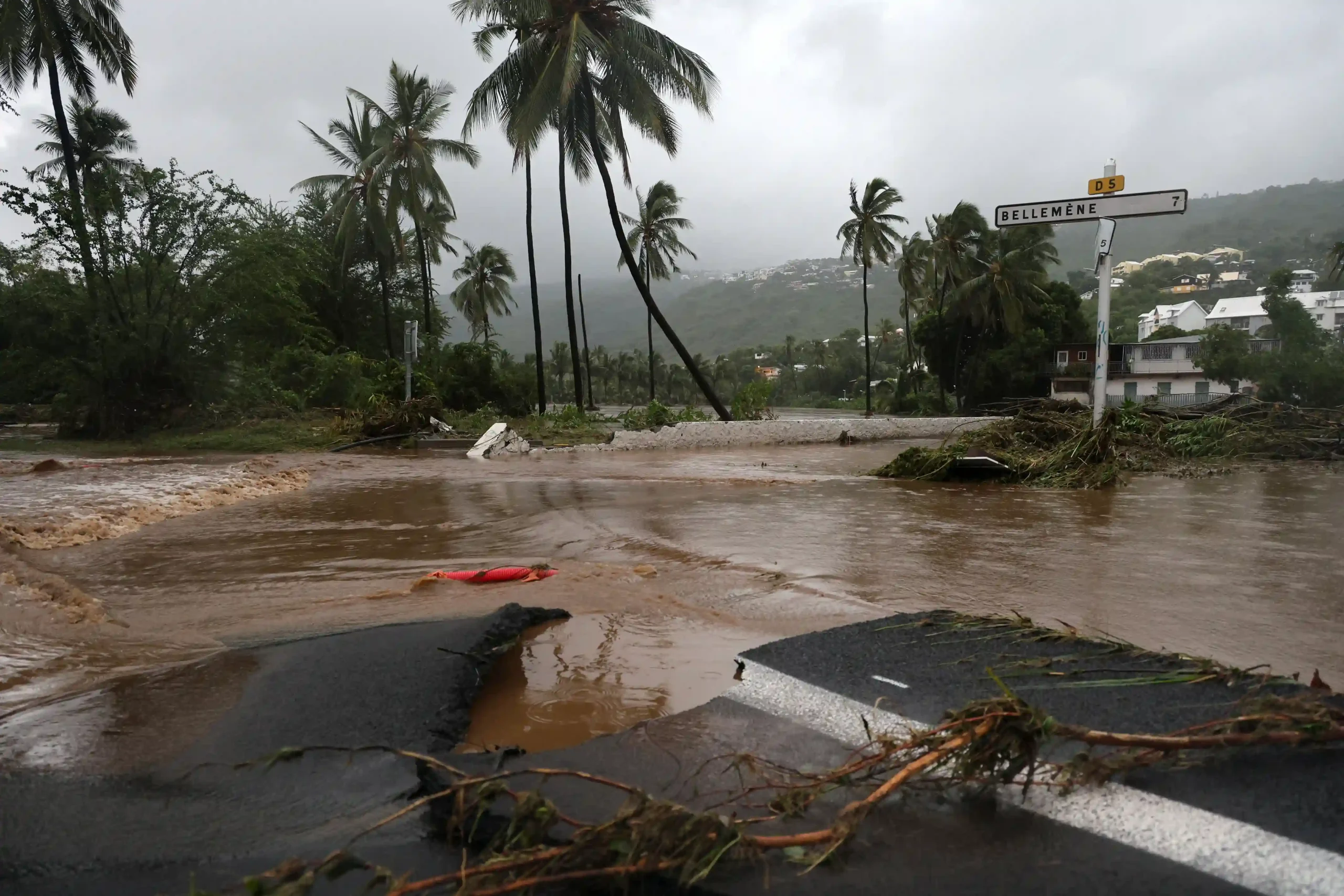 Route inondée bordée de palmiers après le passage d’un cyclone, avec débris et signalisation tordue.
