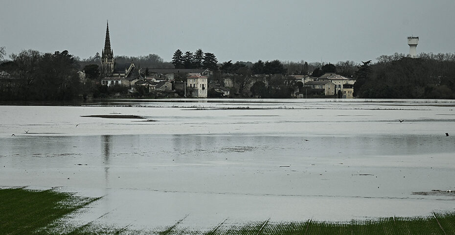 Village en arrière-plan avec une église et des maisons, devant une plaine inondée sous un ciel gris.