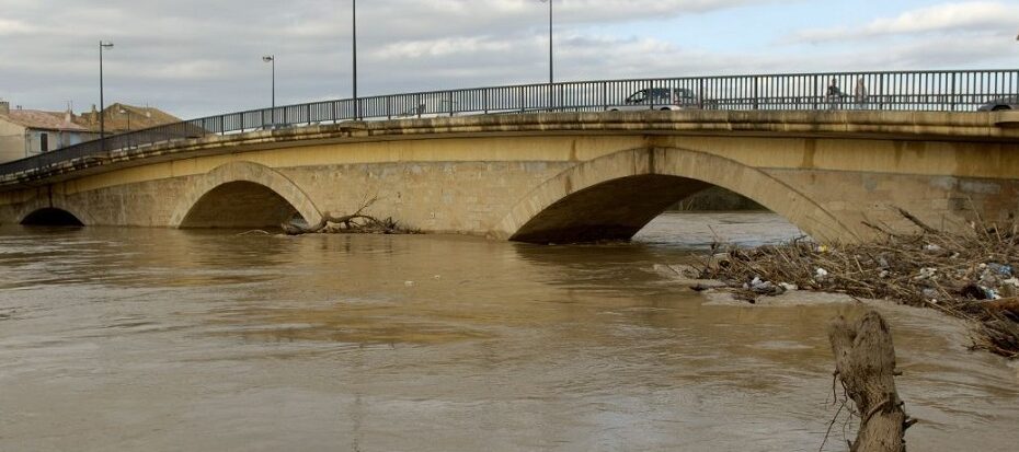 Photographie d’un pont en pierre à deux arches sur une rivière en crue, avec des débris accumulés près des piliers, et des bâtiments en arrière-plan, illustrant un risque d’inondation en Occitanie.