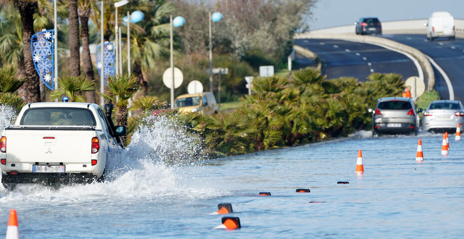 Photographie d’une route partiellement inondée avec trois véhicules traversant l’eau, des cônes de signalisation orange, des palmiers en bordure et une section surélevée sèche en arrière-plan.