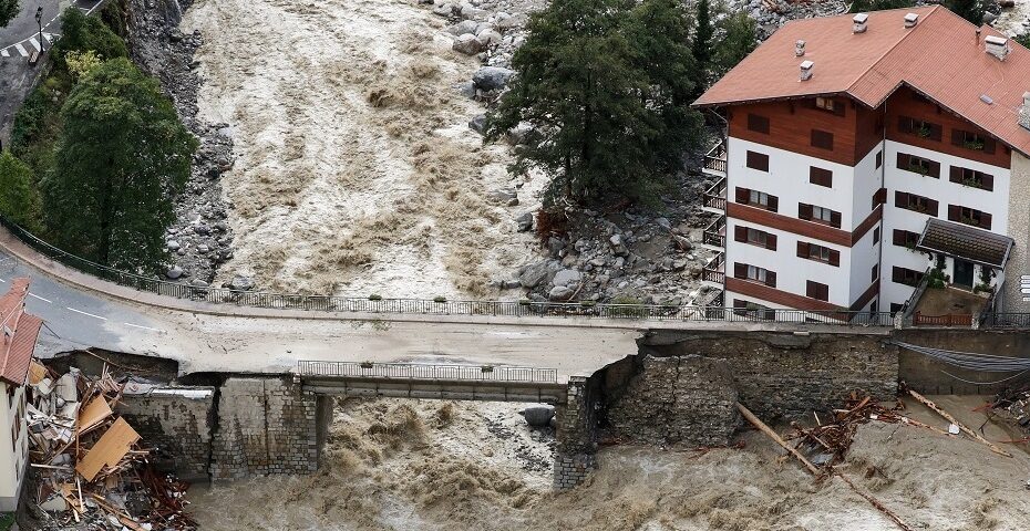 Photographie d’une crue majeure avec une rivière en crue ayant endommagé une route et un pont, des débris visibles et un grand bâtiment à toit rouge situé à proximité de la zone sinistrée.