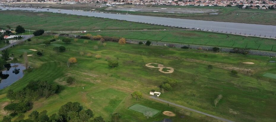 Vue aérienne d’un paysage verdoyant, des plans d’eau, bordé par un canal et quelques bâtiments.
