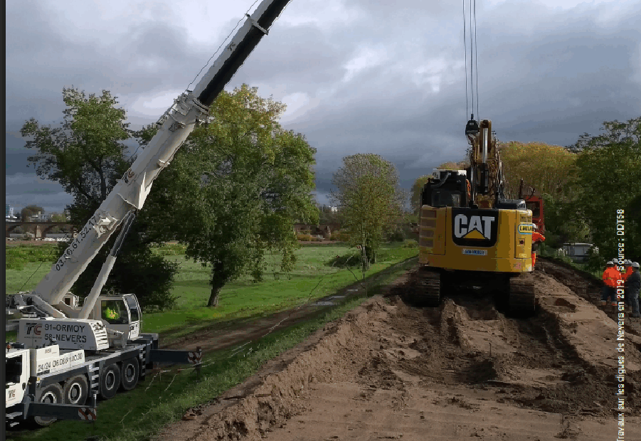 Photographie d’un chantier avec une grue et une pelleteuse intervenant sur une digue ou un remblai, dans le cadre de travaux de prévention contre les inondations, avec des champs et des arbres en arrière-plan.