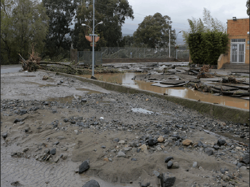 Photographie d’une rue endommagée par une inondation, avec de la boue, des roches et des débris, de l’eau s’écoulant sur le côté, et des arbres et bâtiments en arrière-plan.