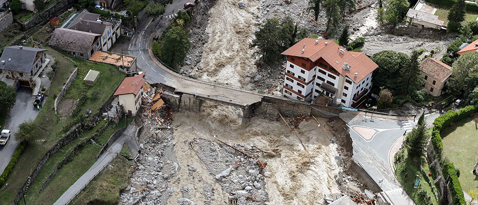 Vue aérienne d’une zone sinistrée par une crue, montrant une route partiellement effondrée, des débris dispersés et plusieurs bâtiments, dont un grand immeuble à toit rouge.