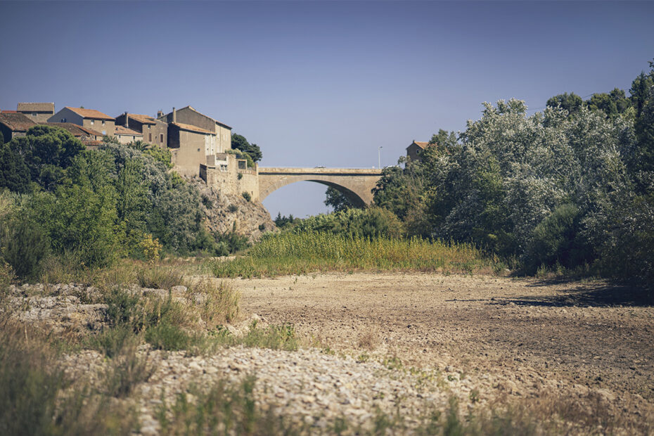 Pont en pierre reliant deux rives, avec un village perché sur la colline et une rivière asséchée au premier plan.